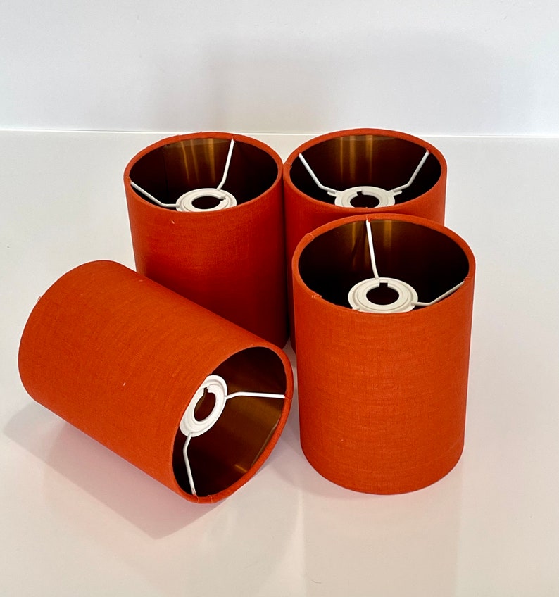 May include: Four orange fabric lampshades with gold interiors. The lampshades are cylindrical and have a white metal frame.