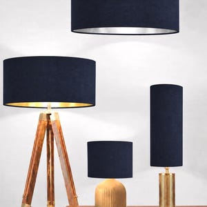 May include: A collection of lighting fixtures featuring navy blue fabric drum shades. Included are a hanging pendant light, a tripod floor lamp with wooden legs, a small table lamp with a ribbed gold base, and a tall table lamp with a gold column base.