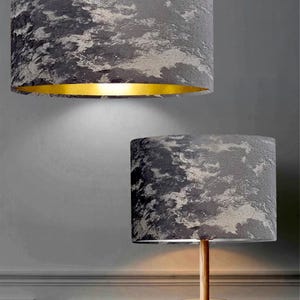 May include: Two cylindrical lampshades with a gray and white abstract pattern. One hangs from the ceiling, the other sits atop a wooden tripod lamp. The interior of the hanging lampshade has a gold finish.