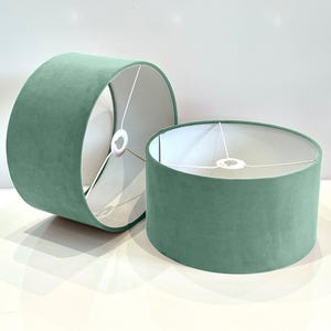 May include: Two teal blue velvet lampshades with white fabric liners. The lampshades are cylindrical and have a smooth, soft texture.