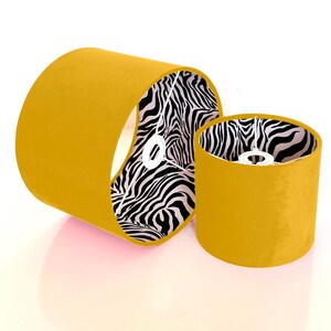 Può includere: Due paralumi color senape con una fodera a stampa zebra in bianco e nero. I paralumi sono cilindrici e hanno una texture di tessuto liscia.