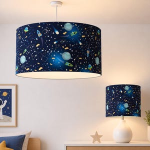 Peut inclure: Un abat-jour bleu foncé et une lampe de table assortie avec un motif sur le thème de l'espace. Le motif comprend des planètes, des fusées et des étoiles. L'abat-jour est cylindrique et la lampe de table a un socle blanc.