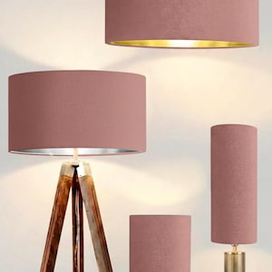 May include: A collection of lighting fixtures featuring dusty rose fabric drum shades. Included are a tripod floor lamp with a wooden base, a pendant light with a gold interior, and two table lamps with gold-toned bases of varying heights.