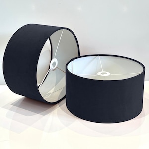 May include: Two black lampshades with white fabric interiors. The lampshades are cylindrical and have a simple design.