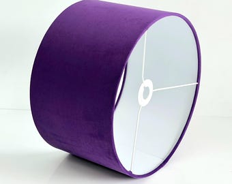 May include: A cylindrical lampshade featuring a deep purple velvet exterior and a smooth white interior lining. The structure is supported by a white metal frame with a central ring fitting, designed to hold the shade securely on a lamp base.