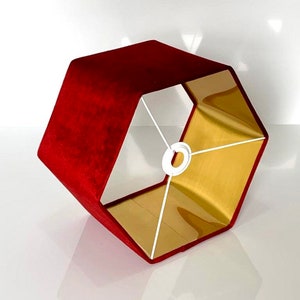 Op de afbeelding: Een zeshoekige lampenkap met een rode fluwelen buitenkant en een gouden binnenkant. De kap heeft een wit metalen frame.