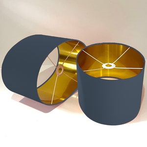 May include: Two oval lampshades with a dark blue fabric exterior and a gold interior. The lampshades are made of fabric and have a metal frame.