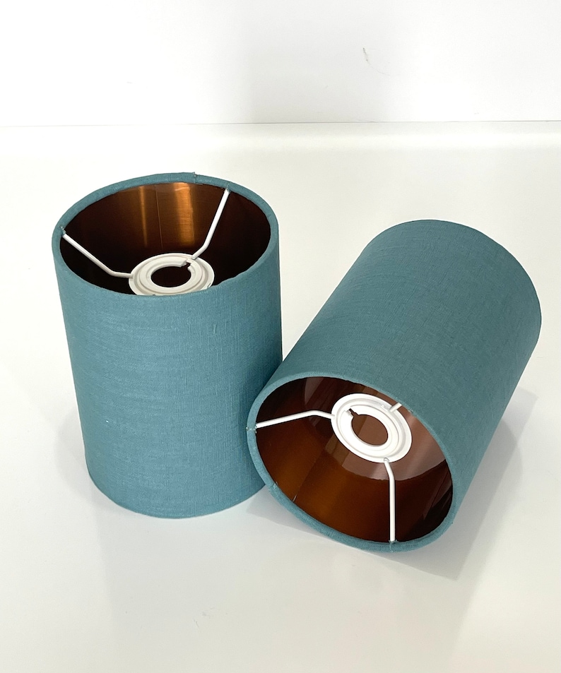 May include: Two teal blue lampshades with a copper lining and white metal frames. The lampshades are made of a textured fabric.