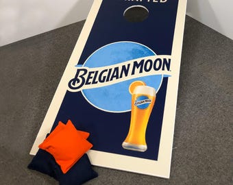 May include: A white wooden cornhole board with a blue and white Belgian Moon beer logo and the text "Artfully Crafted". The board has a hole in the center and is surrounded by two orange bean bags and two blue bean bags.