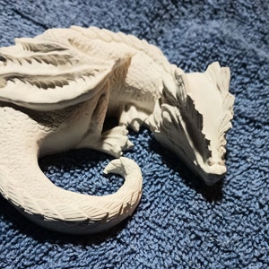 May include: A detailed, white dragon figurine in a resting pose. The sculpture features textured scales, wings, and a curled tail. The dragon is lying on a blue, textured surface.