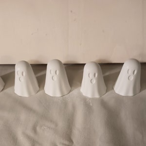May include: Six white ceramic ghost figurines with a simple face design. The ghosts are arranged in a row on a white surface.