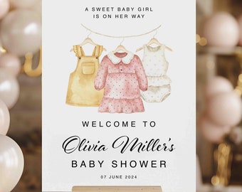 May include: A white sign with a watercolor illustration of three baby clothes hanging on a clothesline. The text reads "A sweet baby girl is on her way. Welcome to Olivia Miller's Baby Shower. 07 June 2024."