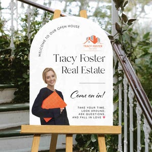 Może przedstawiać: Tablica z informacją o dniu otwartym na drewnianym sztaludze. Na białej tablicy widnieje napis "WELCOME TO OUR OPEN HOUSE" oraz "Tracy Foster Real Estate". Kobieta trzyma pomarańczową teczkę. Na tablicy widnieje r&oacute;wnież napis "Come on in!"