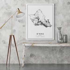 May include: A black and white line art map of Oahu, Hawaii, with the text "O'AHU HAWAII 21.4228&deg;N - 158.0051&deg;W" printed below the map. The map is framed and hanging on a white wall above a whitewashed wooden table.