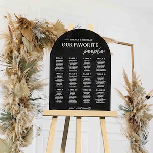 May include: Black acrylic arch-shaped wedding seating chart with white text. The chart lists guest names by table number. The text at the top reads "Harper & Micheal Our Favorite People".