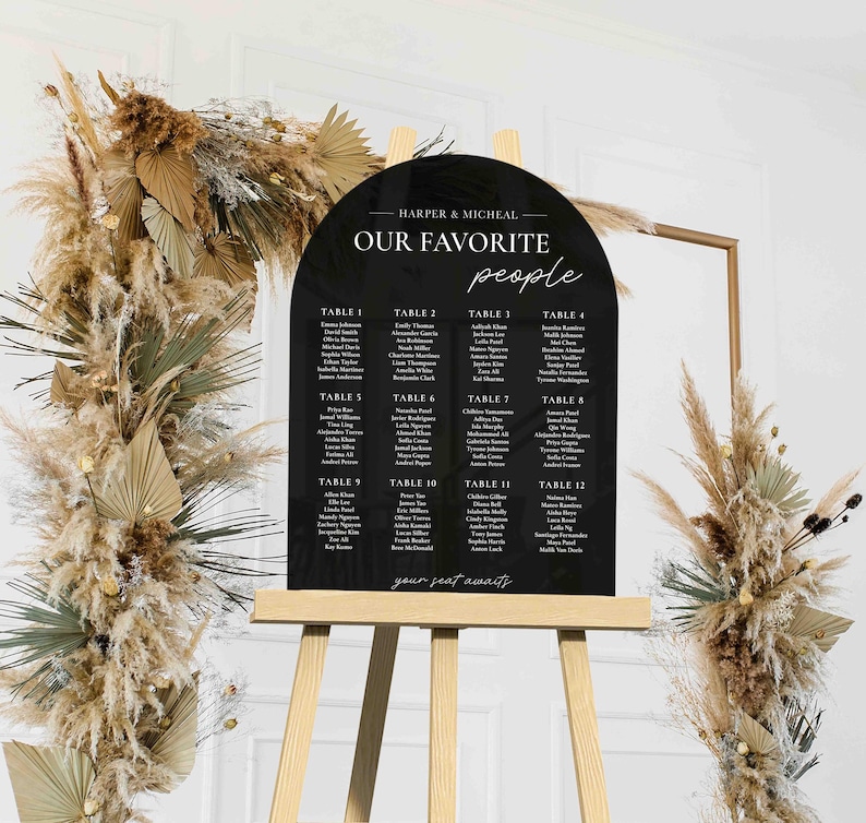 May include: Black acrylic arch-shaped wedding seating chart with white text. The chart lists guest names by table number. The text at the top reads "Harper & Micheal Our Favorite People".
