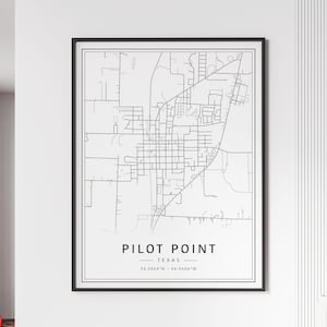 May include: A framed black and white map print of Pilot Point, Texas. The map features a detailed street layout, with the city name and coordinates displayed below. The print is encased in a black frame and mounted on a white wall.