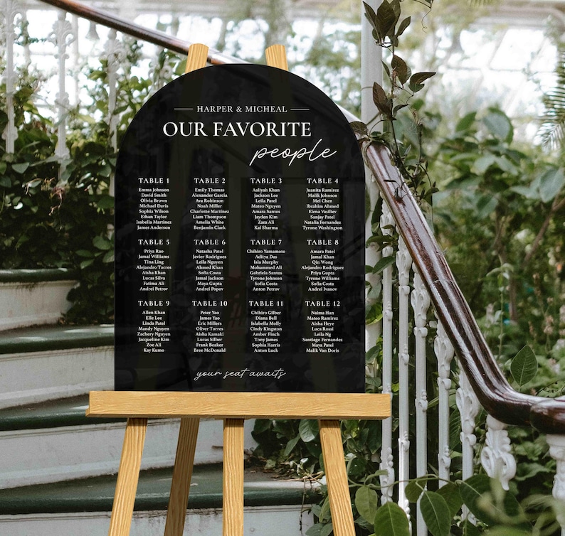 May include: Black arch-shaped acrylic sign with white text listing table numbers and guest names for a wedding reception. The sign reads "Harper & Micheal Our Favorite People" and "Your Seat Awaits".