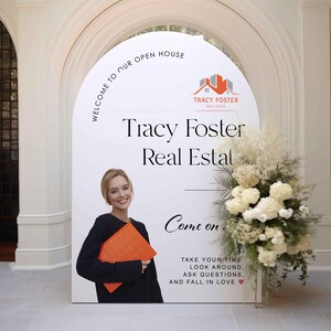 Może przedstawiać: Duży biały znak z napisem "WELCOME TO OUR OPEN HOUSE" i "Tracy Foster Real Estate". Kobieta trzyma pomarańczową teczkę. Znak jest ozdobiony kwiatami i zielenią.