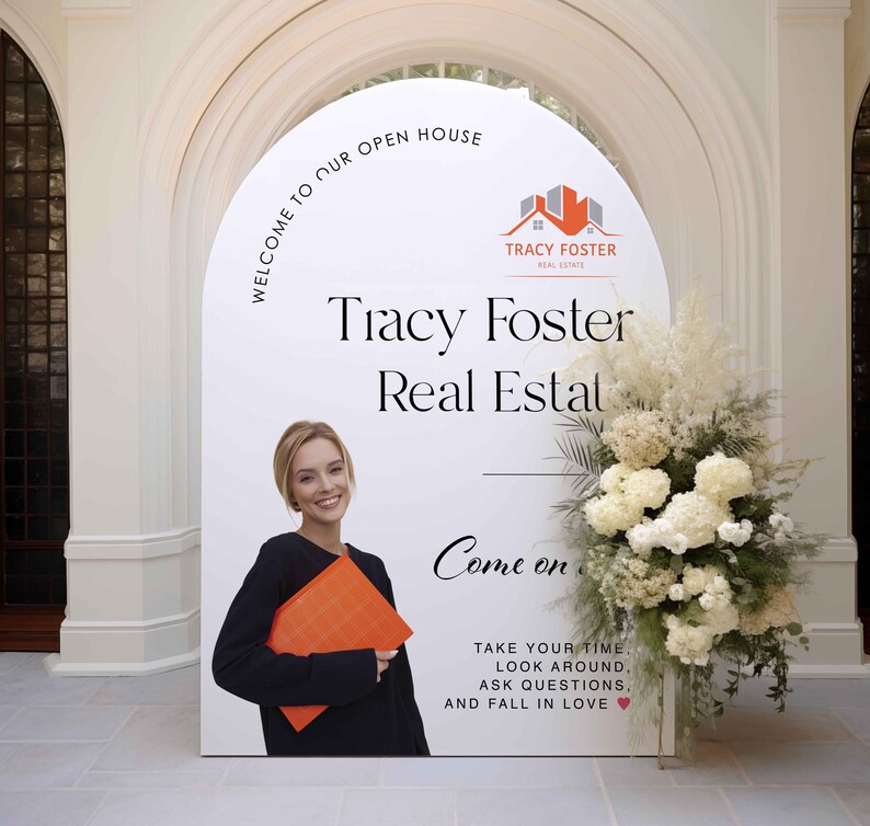 Może przedstawiać: Duży biały znak z napisem "WELCOME TO OUR OPEN HOUSE" i "Tracy Foster Real Estate". Kobieta trzyma pomarańczową teczkę. Znak jest ozdobiony kwiatami i zielenią.