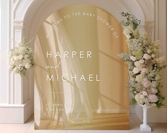 May include: A gold archway with a white background and text that reads "Welcome to the baby shower of Harper and Michael 17 April 2025 Chicago, IL". The archway is decorated with white flowers.