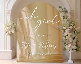 May include: A gold archway with a sheer fabric backdrop and the text "oh girl" in white script. The text "Welcome to Olivia Miller's Baby Shower 06/07/2024" is also in white script.