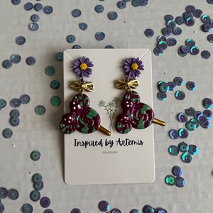 May include: Purple and green swirl earrings with a gold bow and flower charm. The earrings are handmade and the card reads "Inspired by Artemis".
