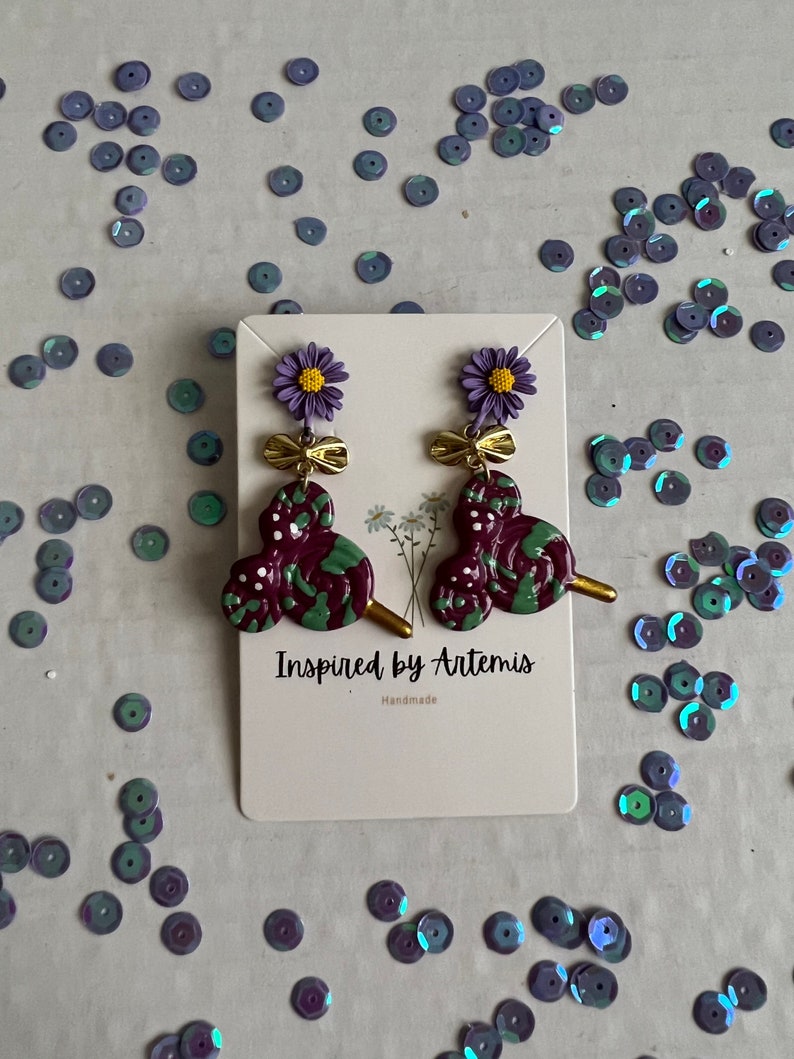May include: Purple and green swirl earrings with a gold bow and flower charm. The earrings are handmade and the card reads "Inspired by Artemis".