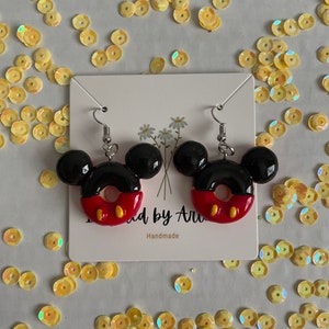 May include: A pair of black and red Mickey Mouse donut earrings with silver hooks. The earrings are handmade and have the text "Made by Ari" and "Handmade" on the card they are displayed on.