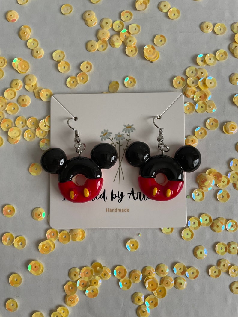 May include: A pair of black and red Mickey Mouse donut earrings with silver hooks. The earrings are handmade and have the text "Made by Ari" and "Handmade" on the card they are displayed on.