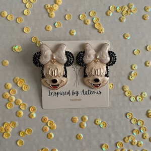 Puede incluir: Un par de pendientes hechos a mano con un personaje de dibujos animados con un lazo blanco, ojos negros y una boca roja. Los pendientes est&aacute;n en una tarjeta blanca con el texto "Inspired by Artemis" y "Handmade".