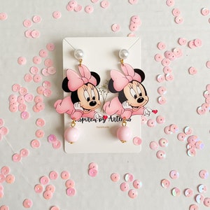 May include: A pair of pink Minnie Mouse dangle earrings with pearl accents. The earrings are handmade and feature a pink bow and a pink pearl dangle.