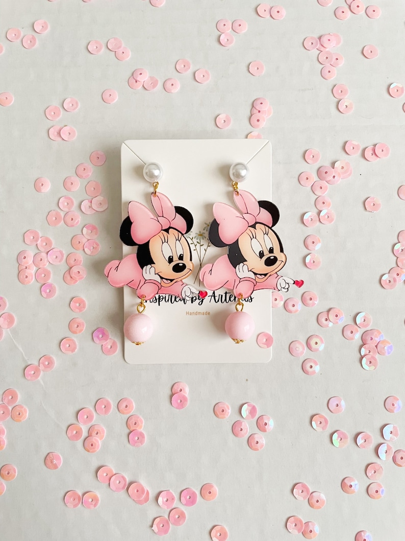 May include: A pair of pink Minnie Mouse dangle earrings with pearl accents. The earrings are handmade and feature a pink bow and a pink pearl dangle.