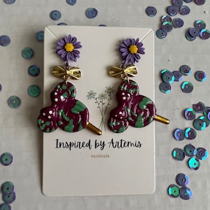 May include: Purple and green swirl lollipop earrings with a purple flower and gold bow charm. The earrings are handmade and the card reads "Inspired by Artemis, Handmade".