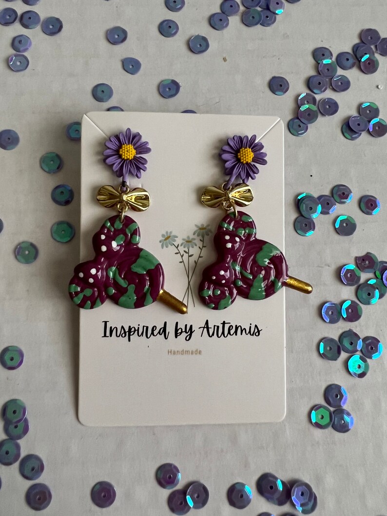 May include: Purple and green swirl lollipop earrings with a purple flower and gold bow charm. The earrings are handmade and the card reads "Inspired by Artemis, Handmade".