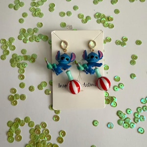 May include: A pair of blue Stitch earrings with red and white striped lollipop accents. The earrings have gold-colored studs and are displayed on a white card with the text "Inspired by Artemis". Green sequins are scattered around the earrings.
