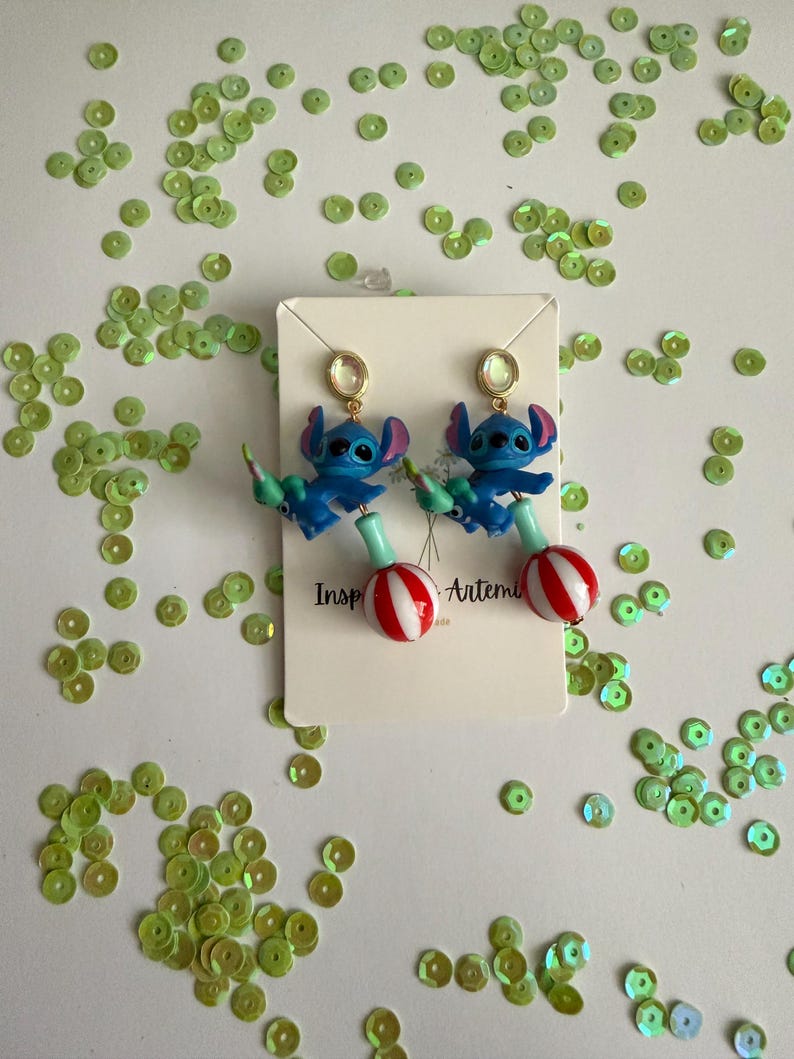 May include: A pair of blue Stitch earrings with red and white striped lollipop accents. The earrings have gold-colored studs and are displayed on a white card with the text "Inspired by Artemis". Green sequins are scattered around the earrings.