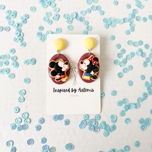 Puede incluir: Un par de pendientes colgantes rojos y marrones con una imagen de dibujos animados de Mickey Mouse y Minnie Mouse. Los pendientes est&aacute;n en una tarjeta blanca con el texto "Inspired by Artemis Handmade".