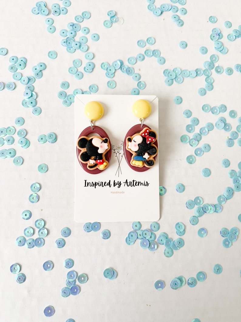 Puede incluir: Un par de pendientes colgantes rojos y marrones con una imagen de dibujos animados de Mickey Mouse y Minnie Mouse. Los pendientes est&aacute;n en una tarjeta blanca con el texto "Inspired by Artemis Handmade".
