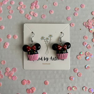 May include: A pair of black and pink cupcake earrings with a white bow and red polka dots. The earrings are on a white card with the text "Inspired by Artemis" and "Handmade".