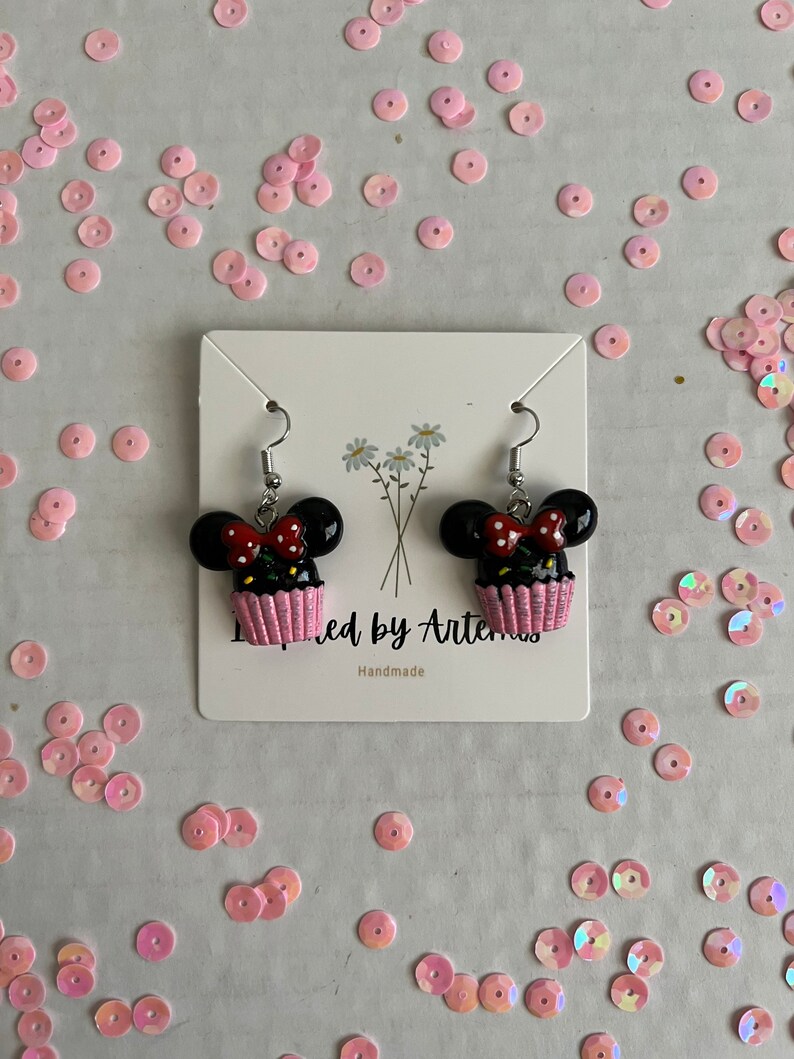 May include: A pair of black and pink cupcake earrings with a white bow and red polka dots. The earrings are on a white card with the text "Inspired by Artemis" and "Handmade".