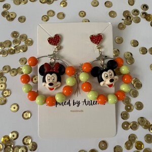 May include: A pair of dangle earrings featuring a cartoon character with a red bow and a heart charm. The earrings are made with orange and light green beads.