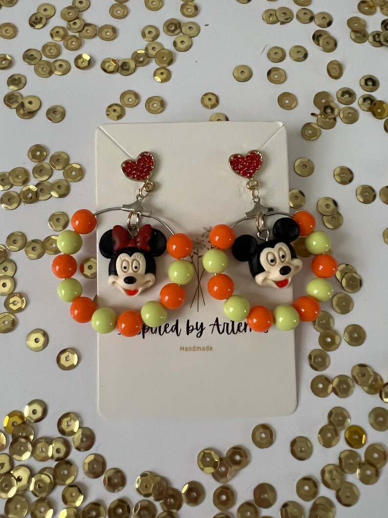 May include: A pair of dangle earrings featuring a cartoon character with a red bow and a heart charm. The earrings are made with orange and light green beads.