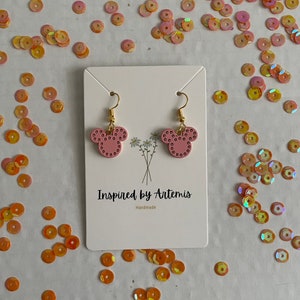 May include: A pair of pink Mickey Mouse-shaped earrings with gold accents. The earrings are displayed on a white card with the text "Inspired by Artemis" and "Handmade".