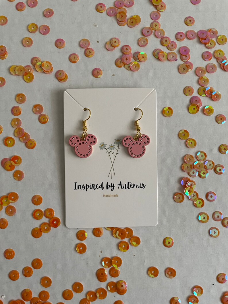 May include: A pair of pink Mickey Mouse-shaped earrings with gold accents. The earrings are displayed on a white card with the text "Inspired by Artemis" and "Handmade".