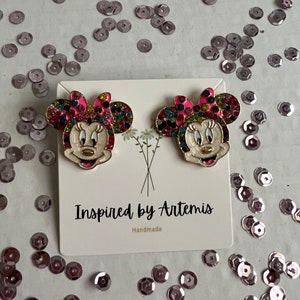 May include: A pair of gold-tone earrings featuring a cartoon character with a pink bow and colorful rhinestones. The earrings are on a white card with the text "Inspired by Artemis Handmade".