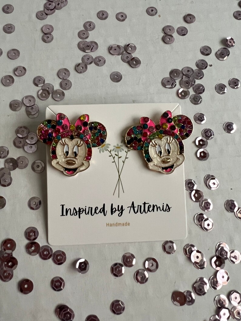 May include: A pair of gold-tone earrings featuring a cartoon character with a pink bow and colorful rhinestones. The earrings are on a white card with the text "Inspired by Artemis Handmade".