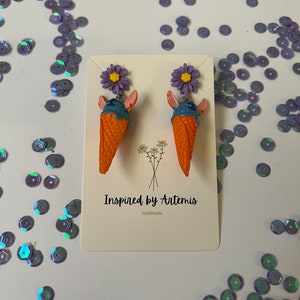 May include: A pair of handmade ice cream cone earrings with purple flowers. The earrings are orange, blue, and purple. The card reads "Inspired by Artemis Handmade".