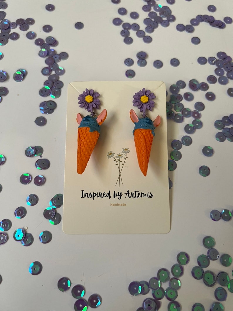 May include: A pair of handmade ice cream cone earrings with purple flowers. The earrings are orange, blue, and purple. The card reads "Inspired by Artemis Handmade".