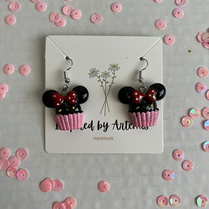 May include: A pair of black and pink cupcake earrings with a red bow and white polka dots. The earrings are handmade and feature a Mickey Mouse silhouette.